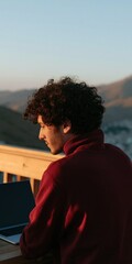 Obraz premium Young man working on a laptop on a balcony at golden hour with mountain view for outdoor lifestyle banner and remote work design