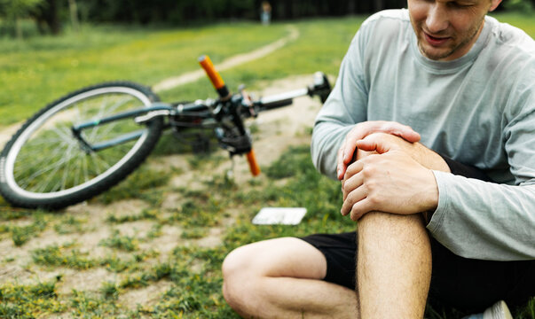 A young Caucasian man sits in a park after falling from a bike, calms the pain in his knee, is a dangerous bike ride in the park on an summer day