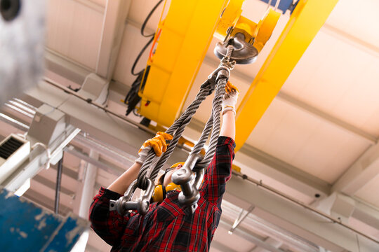 The worker is wearing a hard hat and using a crane to lift a heavy load in a busy factory setting with various equipment and machinery visible in the background.