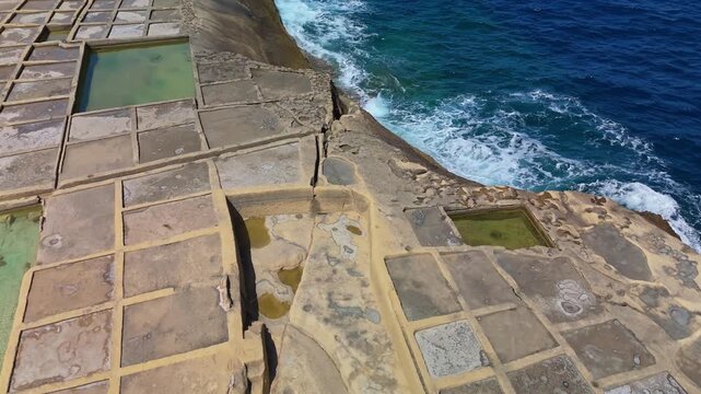 In Xwejni, Żebbuġ on Gozo, Malta, traditional salt pans form rectangular evaporation ponds along the rocky coastline, viewed from a drone overlooking the clear Mediterranean water.
