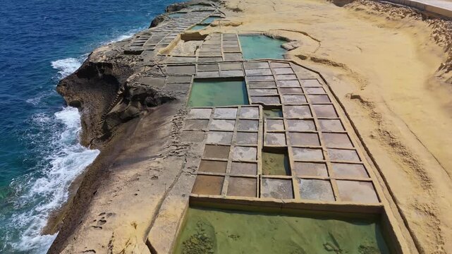 In Xwejni, Żebbuġ on Gozo, Malta, traditional salt pans form geometric rectangular pools along the rocky Mediterranean shoreline in a bird's-eye view under a clear sunny sky.
