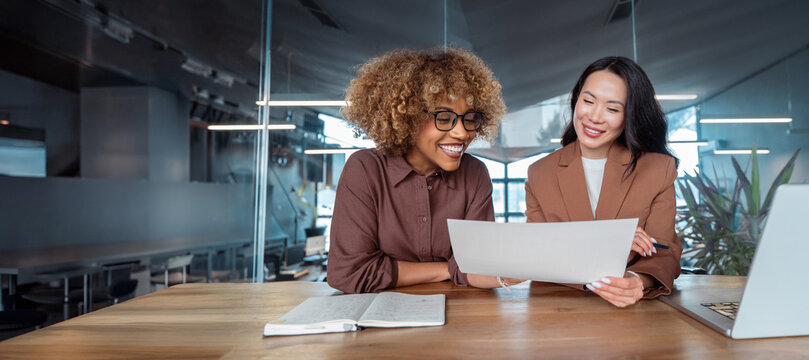 Two smiling businesswomen collaborate at desk in office, reviewing business tax document at laptop, teamwork, mentoring, workplace communication. Smiling multiracial team working. Banner, copy space