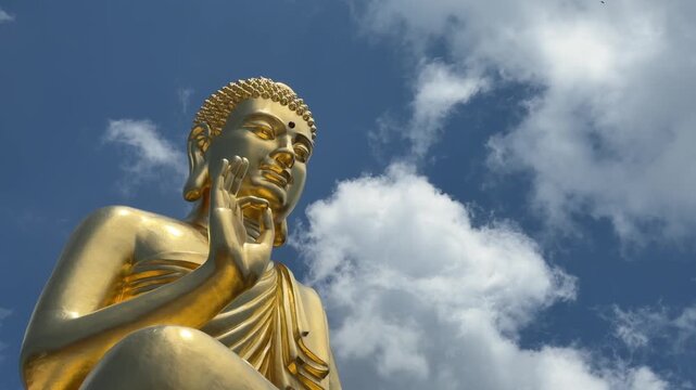 Golden Buddha statue close view with raised hand near face in symbolic mudra against bright clouds. Meditation culture.
