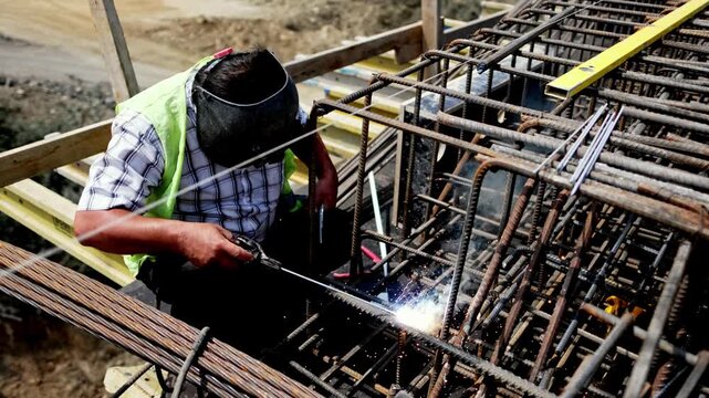 A construction worker welds steel rebar on a bridge structure, showing infrastructure construction, metalwork, safety gear and skilled labor at an outdoor civil engineering site.