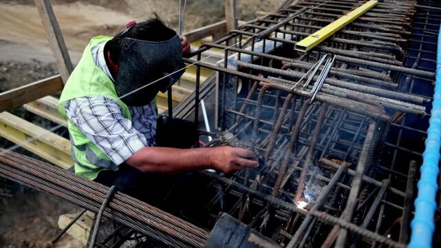 A construction worker welds steel rebar on a bridge structure, showing infrastructure construction, metalwork, safety gear and skilled labor at an outdoor civil engineering site.