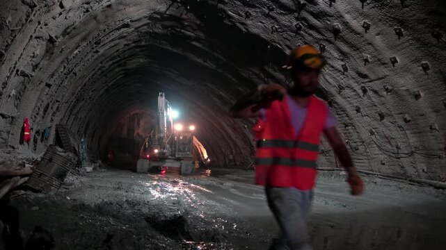 Workers move through a sprayed concrete tunnel with rock bolts, wet ground and machinery lights, showing underground infrastructure construction, safety gear and tunnel engineering progress.