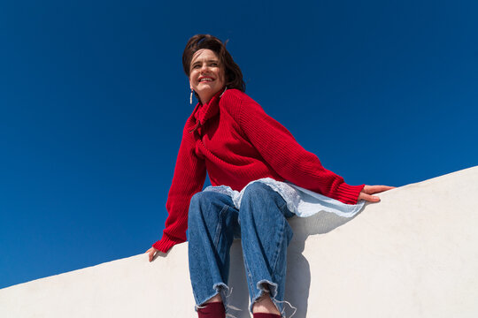 Smiling woman in red sweater sitting on parapet under blue sky