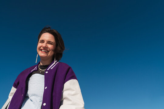 Smiling woman in purple varsity jacket under blue sky
