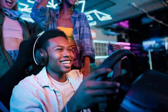 Friends cheer as man plays racing game with steering wheel.