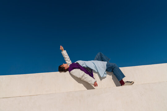 Woman lying on white wall reaching upward against blue sky