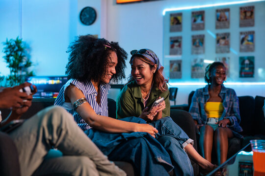 Women laughing over phone while friend plays video game
