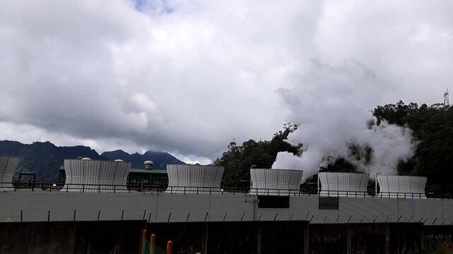 Steam rises from rows of cooling vent towers of an industrial geothermal power plant under cloudy skies. Valencia, Negros Island Region, Philippines