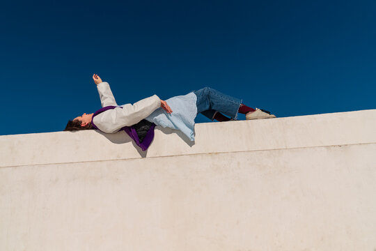 Woman lying on white wall with raised arm under blue sky