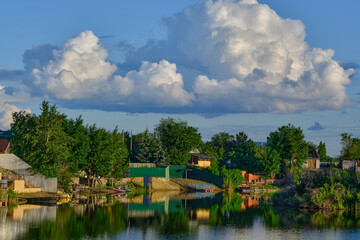 Obraz premium Fluffy cumulus clouds reflect in calm river water near country houses surrounded by green trees.