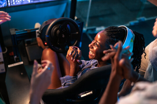 Woman smiling at racing simulator while friends clap and cheer