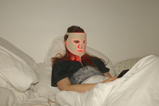 Woman uses Led mask while resting in bed with her phone