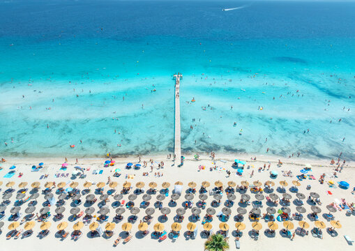 Aerial drone view of Playa de Muro sandy beach in Alcudia, Mallorca, Spain. Wooden pier in turquoise sea water, shore with straw umbrellas, sunbeds, swimming tourists. Summer holiday resort. Top view
