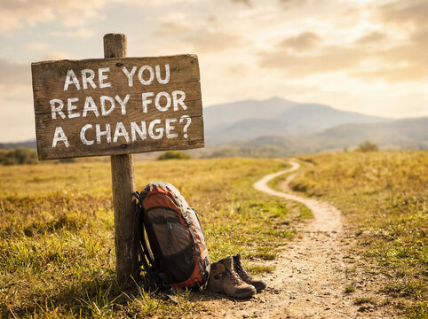 Wooden sign saying are you ready for a change next to a backpack and hiking boots on a winding trail toward mountains