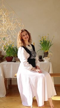 Elegant mature woman wearing embroidered clothing and handcrafted jewelry while sitting in a sunlit interior decorated with spring flowers. Cultural identity, personal style, authentic fashion and fem