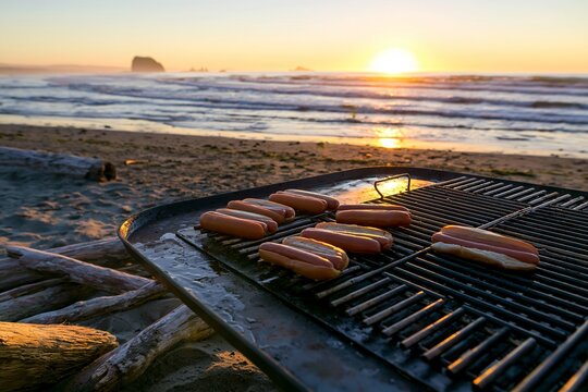 Beachfront barbecue at sunset with hot dogs cooking on portable grill near ocean waves.