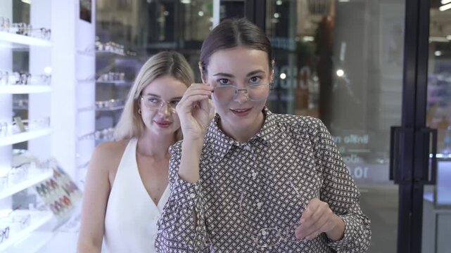 Close-up of a blonde female buyer trying on new designer eyeglasses and checking her look, with a professional female optician standing nearby to assist in the selection.