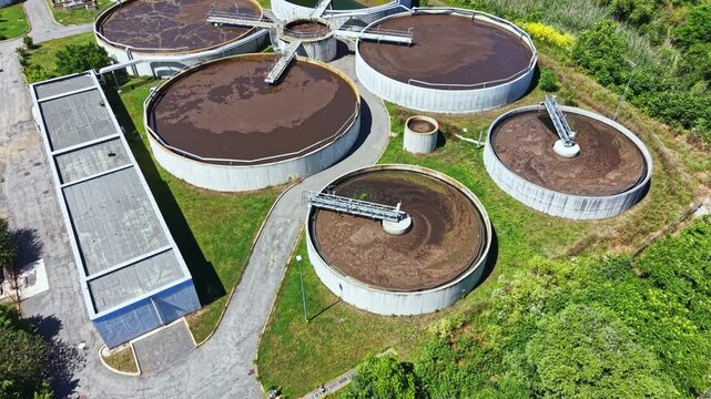 A treatment facility works on processing wastewater using large circular tanks and pipes under a clear sky. Surrounding greenery adds to the scene while machines perform tasks.