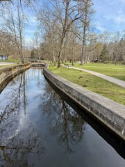 Fototapeta premium Vertical view of a calm stone canal and a small footbridge in a peaceful manor park with leafless trees in spring, Keila-Joa, Estonia