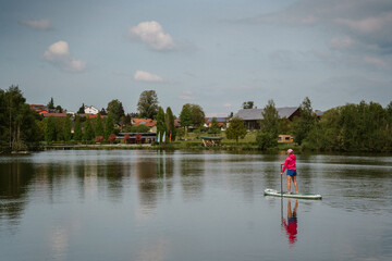 Fototapeta premium Woman paddleboarding on calm lake surrounded by green trees in summer daylight with blue sky. Active outdoor water sports recreation, healthy lifestyle fitness, ecotourism weekend getaway concept