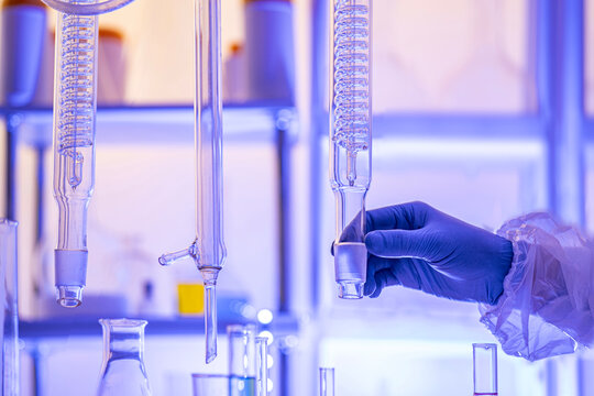 Close up of a gloved hand holding a glass rectification fractionating column among laboratory glassware. Concept for distillation, chemical engineering and analytical chemistry, copy space.