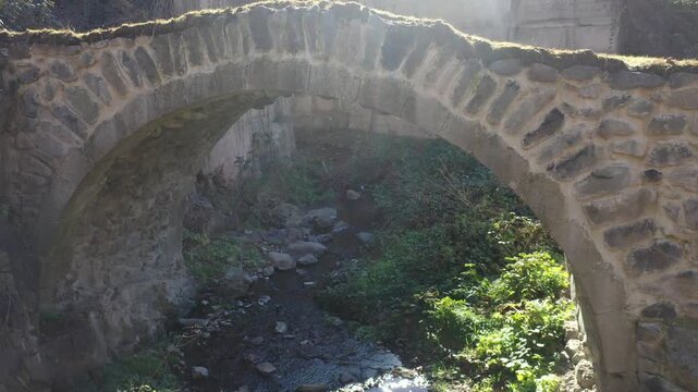 A beautiful historic stone arch bridge stands over a gently flowing river stream surrounded by lush green grass and rocky hillsides on a bright and sunny spring day.

