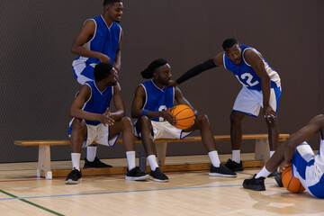 Naklejka premium African American male basketball players in blue jerseys sitting on bench at gym holding basketball
