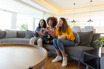 Diverse female friends playing console game on sofa holding controllers and eating from bowl © wavebreak3