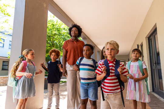 Diverse children standing under covered walkway with African American teacher, adjusting backpacks
