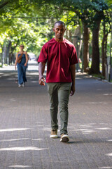 Obraz premium African American man walking on paved path under dappled canopy wearing burgundy shirt, olive pants