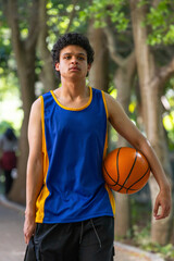 Fototapeta premium African American man holding orange basketball standing on park path in blue jersey, black shorts