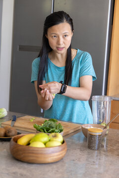 Asian woman in teal shirt smartwatch preparing smoothie in kitchen with cutting board