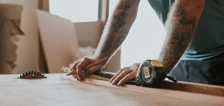 Tattooed man measuring wood at a workshop table with a tape measure