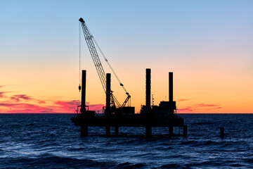 Obraz premium Offshore crane platform silhouette at sunset over calm ocean waters