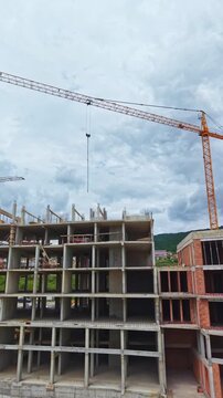 Workers are building a multi-storey building using concrete blocks and steel structures. A crane is lifting materials at the construction site under a cloudy sky.