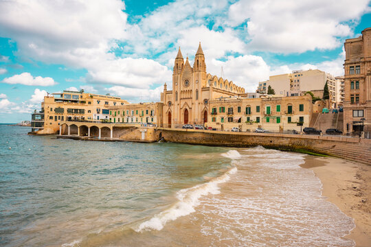 This scene depicts Balluta Bay in St. Julian's, Malta, featuring a beach promenade.The large Neo-Gothic building is the Carmelite Church (Parish Church of Our Lady of Mount Carmel).The bay is a popula