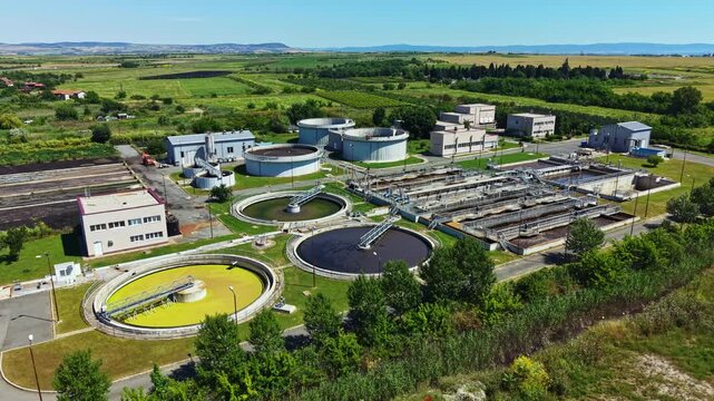 The scene shows a wastewater treatment facility located in a rural area. Various treatment tanks and buildings are present, with green fields in the background.