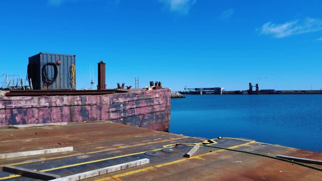 Drone flight above a rusty industrial dock and barge in Matane Harbor, featuring a moored ferry, calm waters, and a clear blue coastal sky.