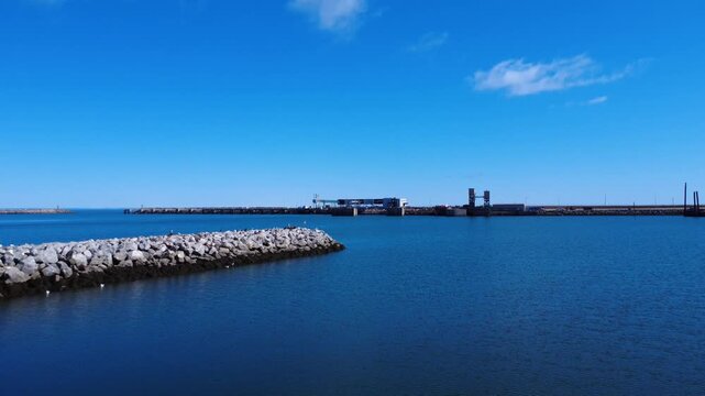 Drone footage of seagulls soaring above a calm blue harbor with a rocky breakwater and distant jetty under a clear sky in Matane, Quebec, Canada.