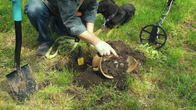 Man with dog digs in garden soil using a trowel, uncovering buried items while a metal detector rests nearby on the grass