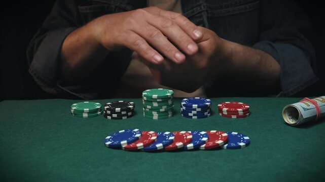 Male player in a denim jacket organizes poker chips and reveals playing cards three aces on a green felt table, with a roll of cash and additional chips positioned nearb