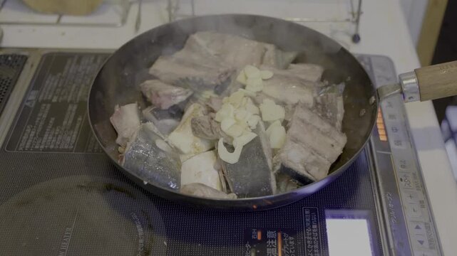鰤アラのオリーブ煮を調理中、焼き霜した鰤アラの上にニンニクを乗せ、フライパンを振り、水を500cc投入するシーン