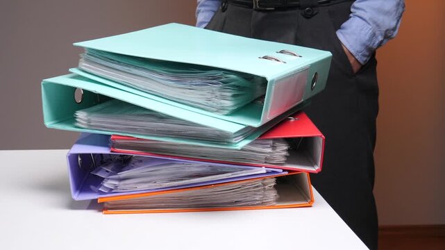 Businessman standing in an office, managing a stack of colorful binders filled with documents, symbolizing organization, administration, and the workload of paperwork