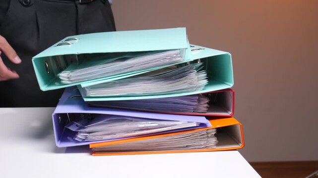 Person's hands holding and stacking colorful ring binders filled with documents and paperwork, representing organization, administration, and bureaucracy in a business office