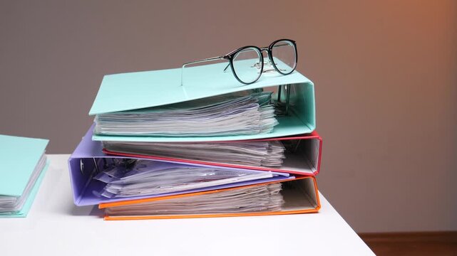Business person reaching for reading glasses placed on a pile of colorful ring binders filled with paper documents, symbolizing organization, management, and diligent office work