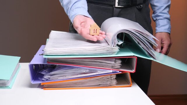 Hand holding small wooden house model over colorful binders and scattered financial paperwork, symbolizing property purchase, mortgage planning, and real estate transaction process